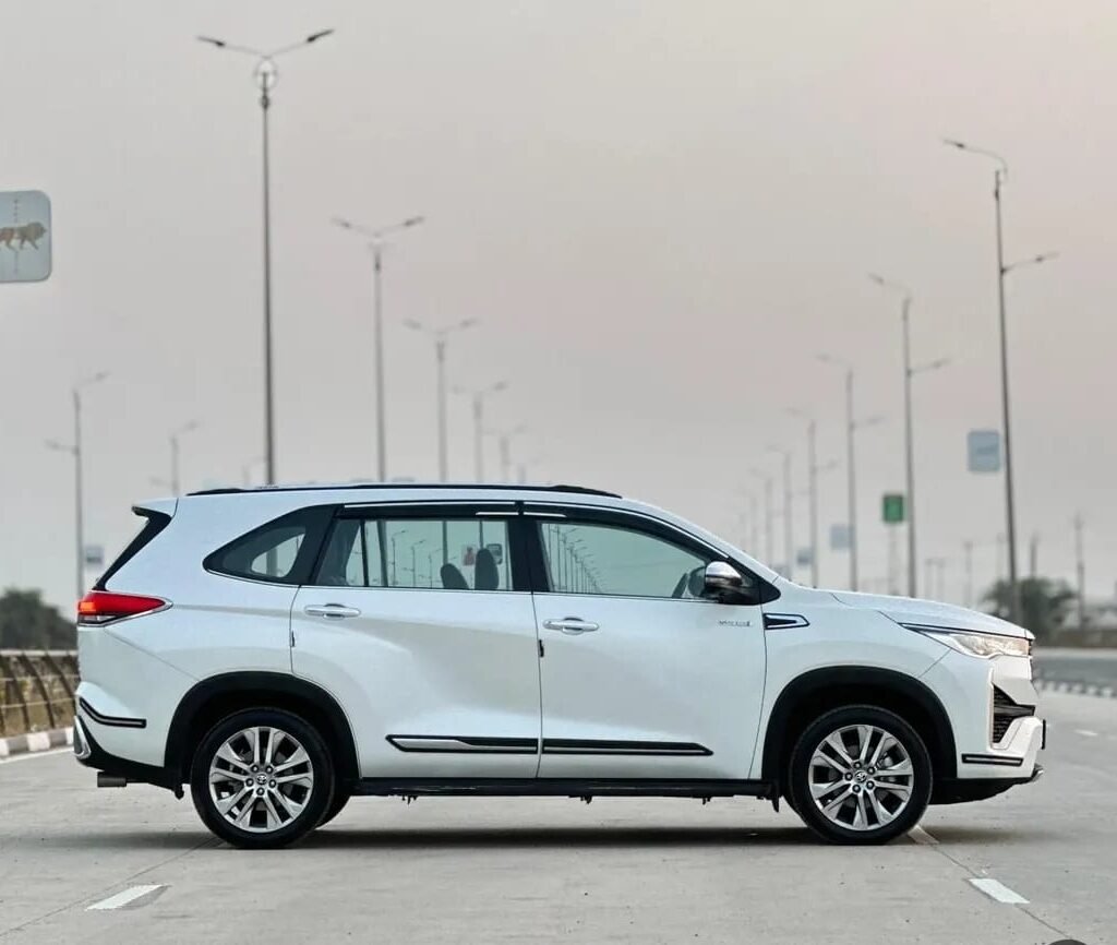 Side profile of a white Toyota Innova Hycross parked on a wide highway, showcasing its premium SUV-inspired design and spacious family-friendly body.