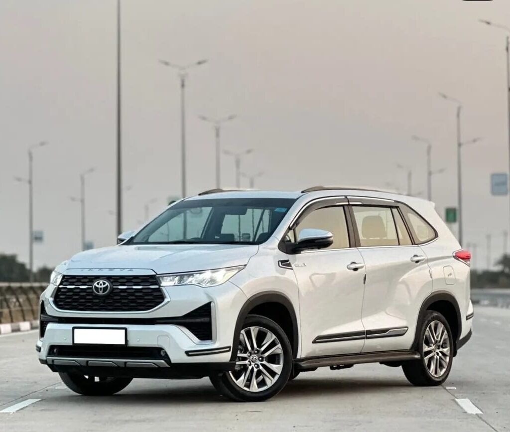 Front three-quarter view of a white Toyota Innova Hycross parked on a modern highway, featuring a bold grille, LED headlights, and premium alloy wheels.
