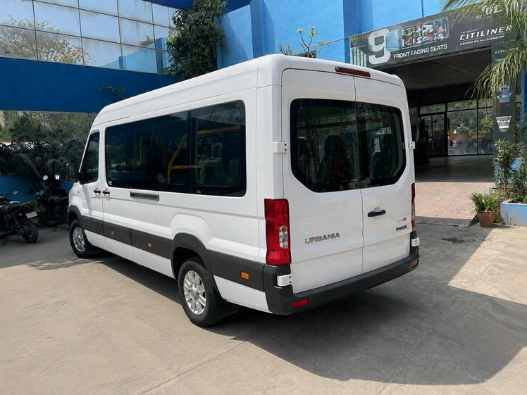 Rear left side view of a white Force Urbania luxury van parked outside a modern commercial building, showcasing its spacious design and premium passenger transport styling.