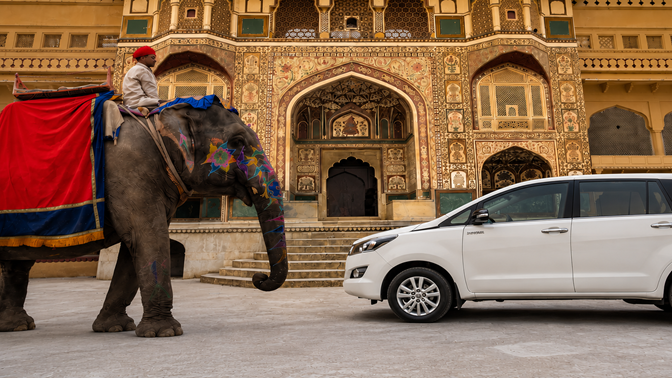 White Toyota Innova Crysta facing a decorated elephant at Amer Fort Jaipur, showcasing Limo Car India's premium sightseeing and luxury taxi service.