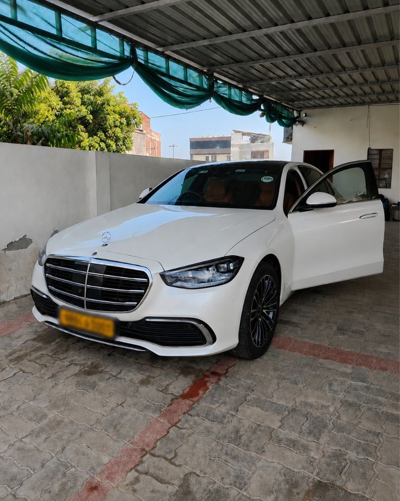 White Mercedes-Benz S-Class luxury car parked under a covered driveway with the rear passenger door open and a blurred yellow number plate.